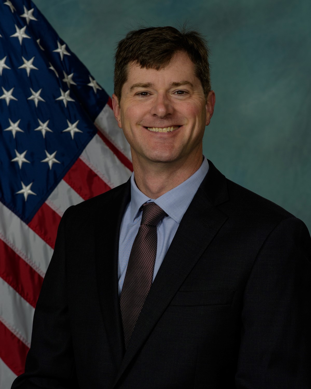 Dan Randolph stands in a suit and tie before a blue background and the American flag