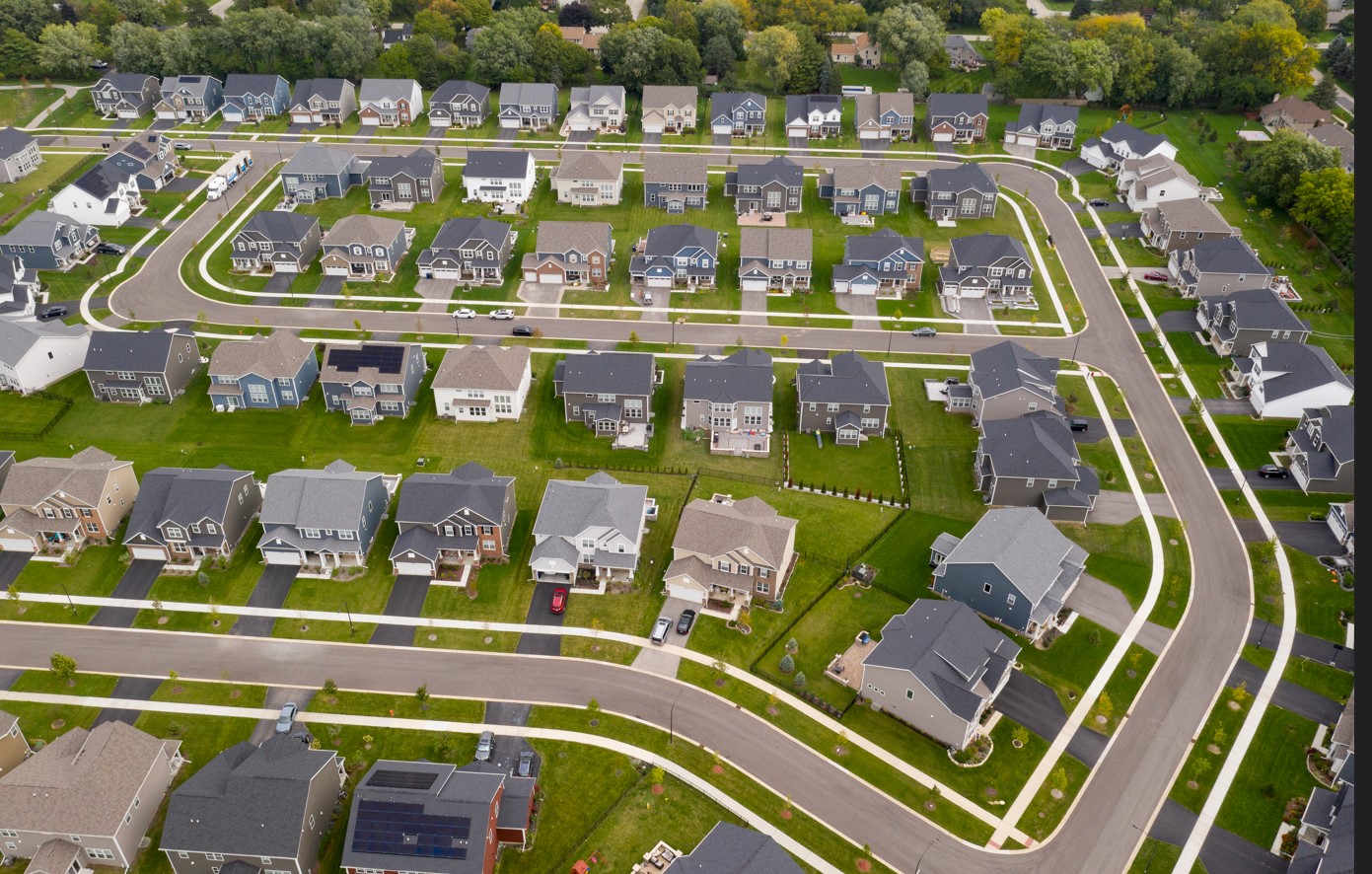 Aerial view of a suburban neighborhood with rows of single-family homes, driveways and curbside street parking along curved residential streets.
