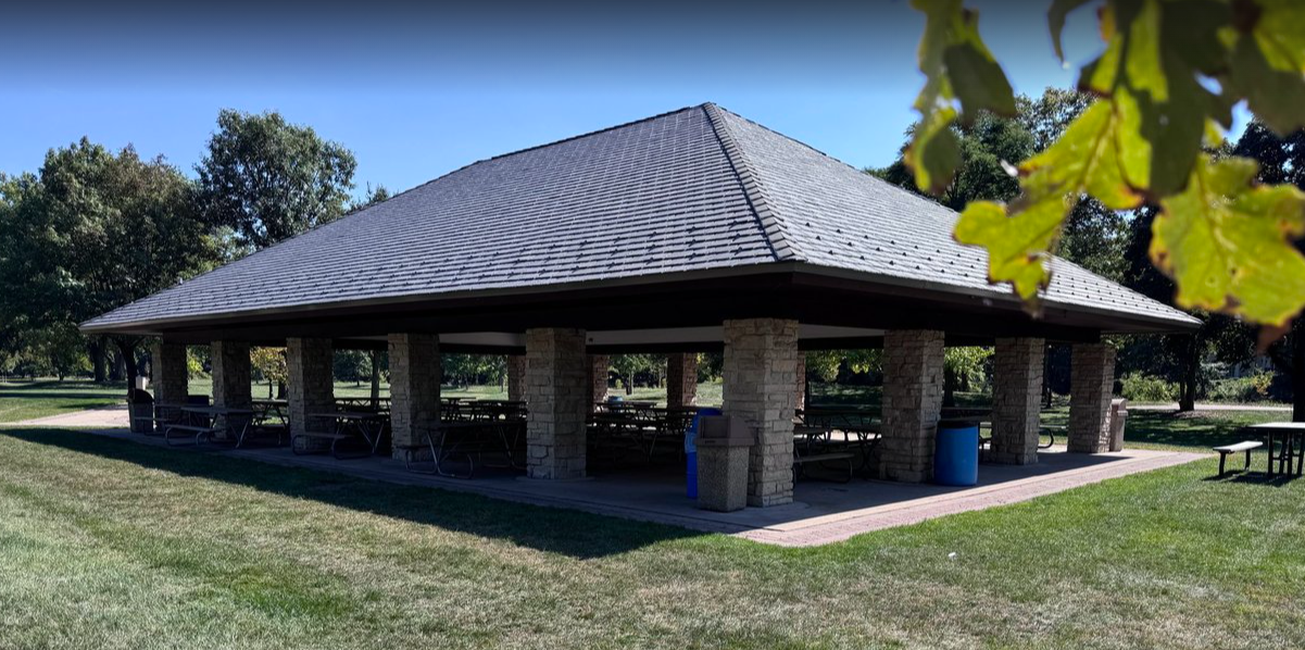 A large, open-air picnic shelter with a gray shingle roof sits in a grassy park. The shelter is supported by numerous stone columns and contains several picnic tables. A clear blue sky is visible above, and trees with green leaves border the area in the background.