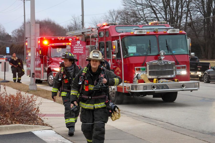 Two City of Naperville firefighters, fully dressed in their gear, stride away from a fire truck and ambulance with lights illuminated.