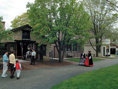 People dressed in period costumes stroll down a pathway with modern-day visitors, as the buildings of Naper Settlement provide a backdrop.