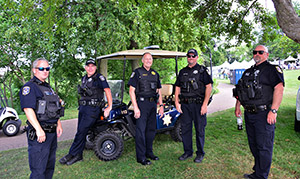 Five officers in uniform stand in front of a police golf cart at an outdoor festival.