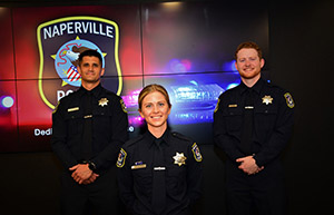 Three officers in uniform smile while standing in front of a screen bearing the NPD patch.